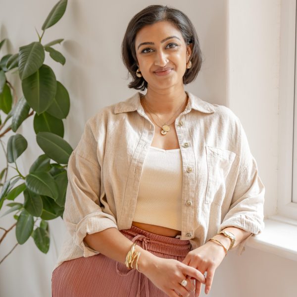 Smiling woman in a neutral outfit standing by a window with a plant in a bright, modern interior.